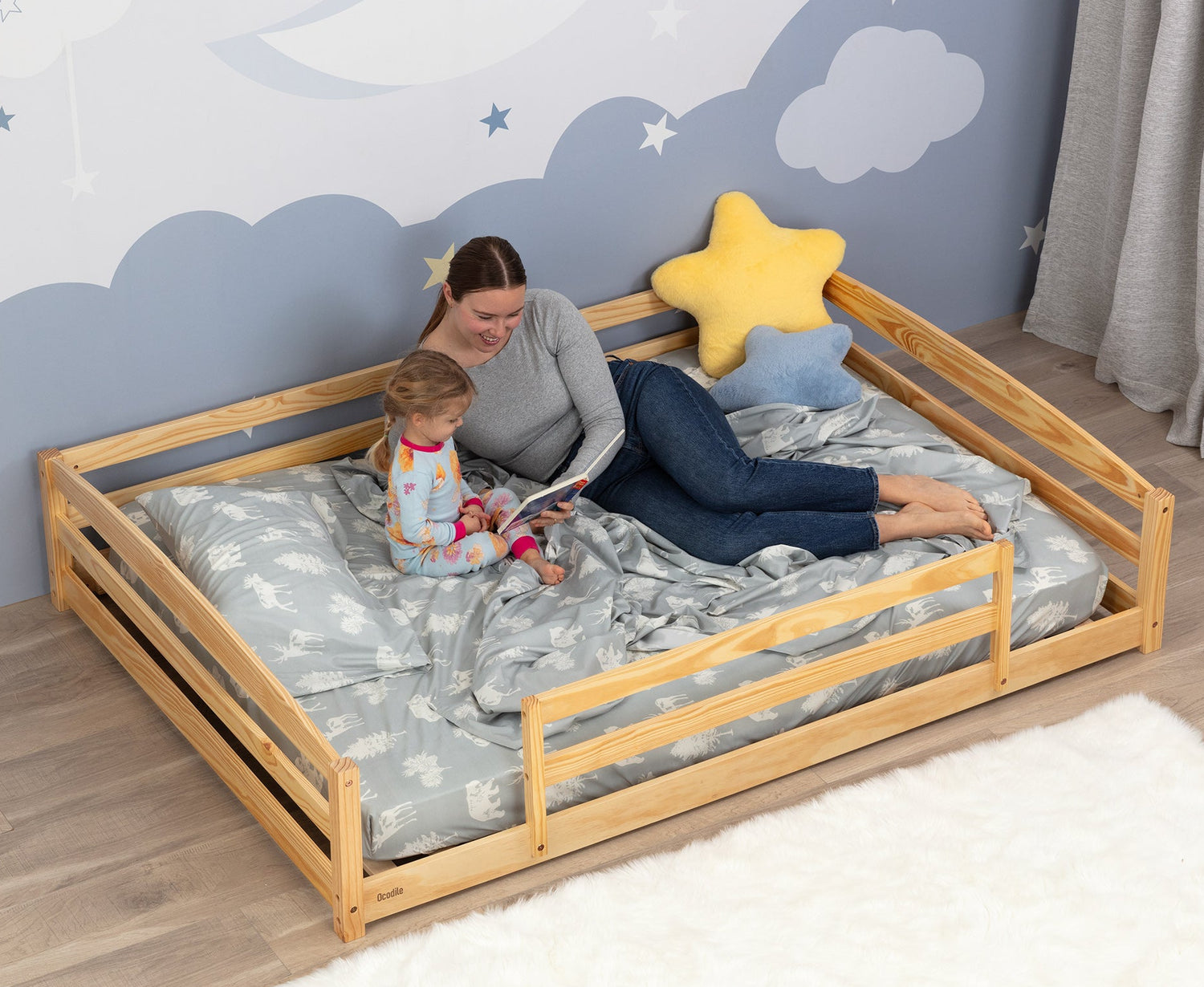 Woman and child on a floor wooden bed with star-shaped pillow in a room with cloud and moon wall decor.